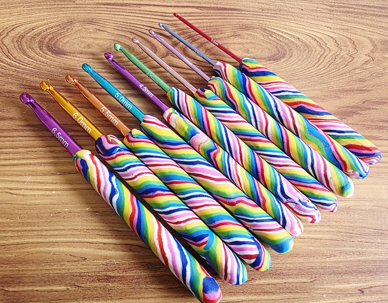 May include: A set of nine crochet hooks with colourful, swirled, multi-coloured handles. Each metal hook features a specific size printed on the shaft, ranging from 2.5mm to 6.5mm. The hooks are arranged in a diagonal row, showcasing various metallic colours.