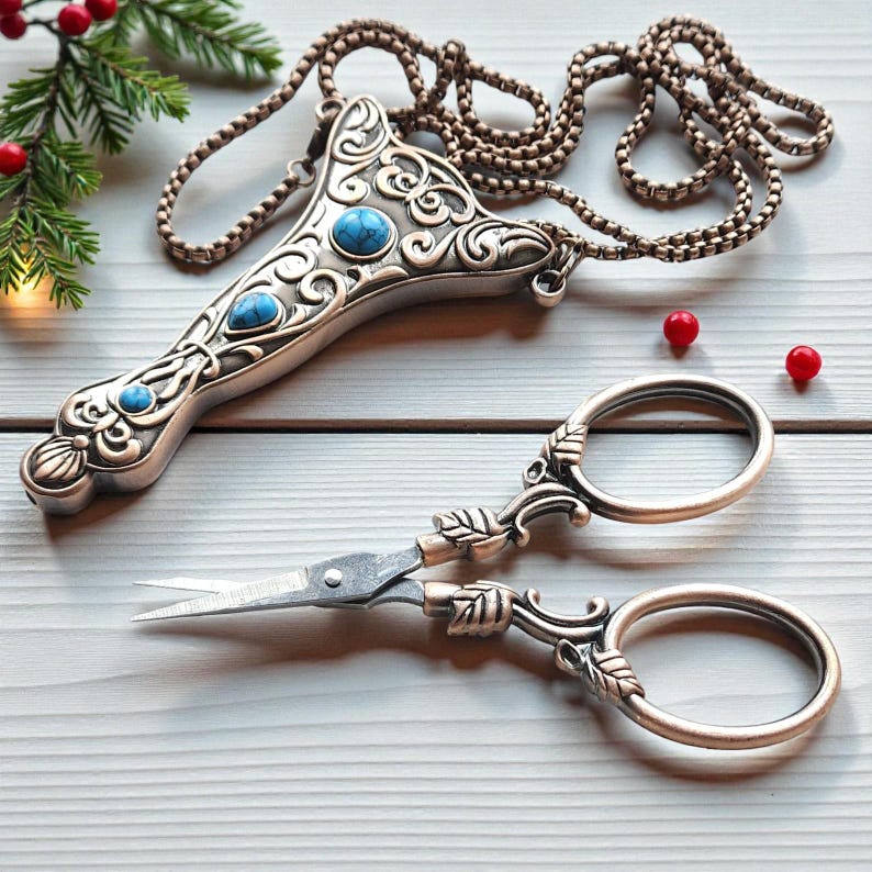 May include: An ornate silver pendant with turquoise detailing and a pair of decorative scissors. The scissors feature a leaf design on the handles. The pendant is attached to a chain. The scissors and pendant are on a light wooden surface.