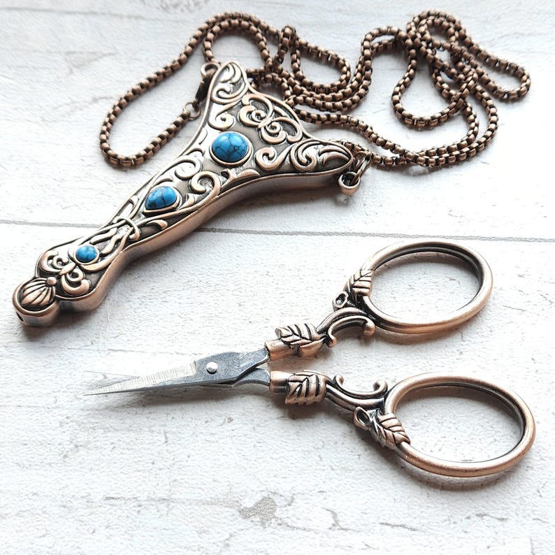 May include: A copper-toned pendant with turquoise-coloured stones and a pair of small scissors. The pendant has an ornate design and a chain. The scissors feature leaf-shaped details on the handles, creating a vintage aesthetic.