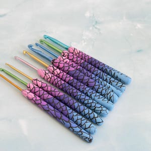May include: A set of seven crochet hooks with metallic tips in gold, green, and blue. Each handle features a gradient transition from pink to purple to blue, overlaid with a black crackle pattern that resembles scales or a textured surface.