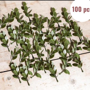May include: A collection of 100 artificial green leaf sprigs, each with multiple oval-shaped leaves. The sprigs are arranged on a light-colored wooden surface. The text "100 pcs" is in a white circle in the upper right corner.