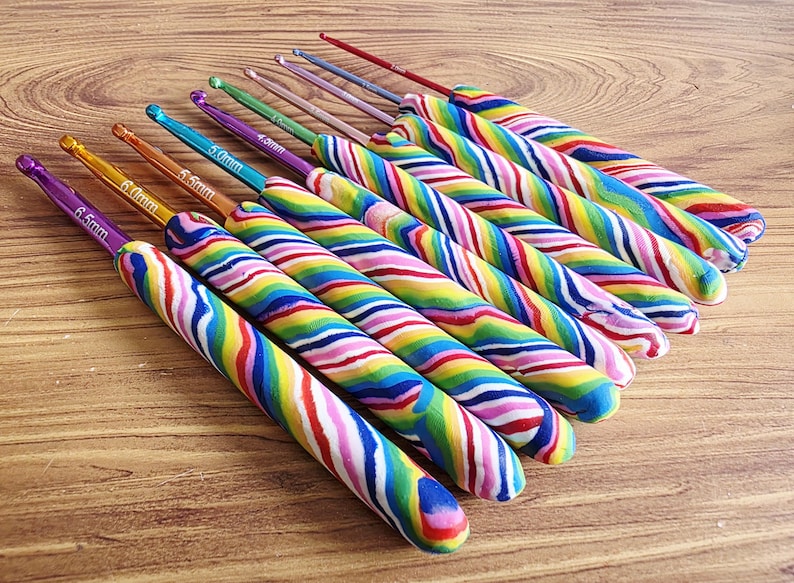 May include: A set of nine crochet hooks with colourful, swirled polymer clay handles and metallic tips. Each hook features a size marking ranging from 2.0mm to 6.5mm printed on the metal shaft. The handles display a vibrant, multi-coloured striped pattern.