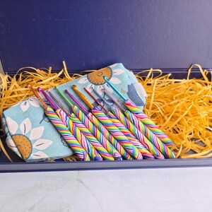 May include: A set of crochet hooks with rainbow-striped handles arranged in a light blue fabric pouch featuring a sunflower pattern, all contained within a dark blue rectangular gift box filled with yellow shredded paper filler.