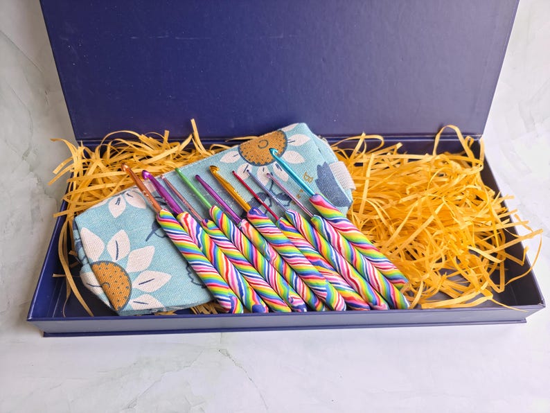 May include: A set of crochet hooks with rainbow-striped handles arranged in a light blue fabric pouch featuring a sunflower pattern, all contained within a dark blue rectangular gift box filled with yellow shredded paper filler.