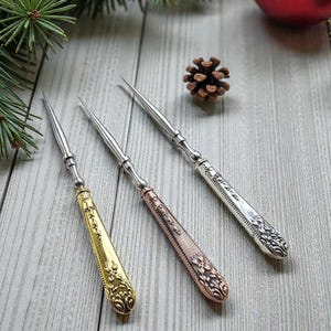 May include: Three decorative silver and gold-toned cheese knives with ornate handles. The handles are gold, copper, and silver, with intricate floral designs. The knives are arranged on a light wood surface, with pine needles and a pine cone in the background.