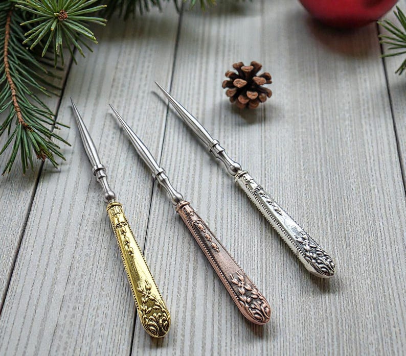 May include: Three decorative silver and gold-toned cheese knives with ornate handles. The handles are gold, copper, and silver, with intricate floral designs. The knives are arranged on a light wood surface, with pine needles and a pine cone in the background.