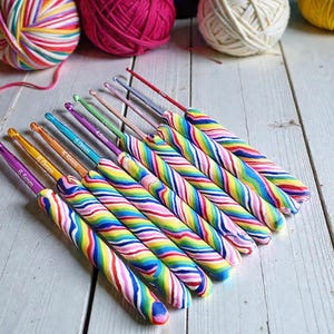 May include: A set of nine crochet hooks with colorful, swirled, rainbow-patterned handles. Each metal hook features a specific size printed on the shaft, ranging from 6.5mm down to smaller sizes, arranged in a diagonal row on a white wooden surface.