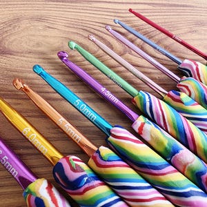 May include: A set of nine metal crochet hooks with colourful, swirled polymer clay handles. Each hook features a distinct metallic colour and a printed size ranging from 2.0mm to 6.5mm, including 2.5mm, 3.0mm, 3.5mm, 4.0mm, 4.5mm, 5.0mm, 5.5mm, and 6.0mm.