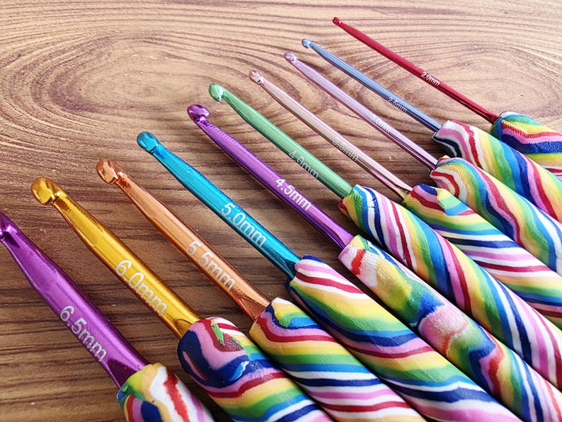 May include: A set of nine metal crochet hooks with colourful, swirled polymer clay handles. Each hook features a distinct metallic colour and a printed size ranging from 2.0mm to 6.5mm, including 2.5mm, 3.0mm, 3.5mm, 4.0mm, 4.5mm, 5.0mm, 5.5mm, and 6.0mm.