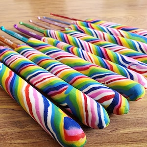 May include: A set of crochet hooks featuring metallic tips in various colours and thick, ergonomic handles with a swirling, multi-coloured rainbow pattern. Each hook displays its size clearly on the metal shaft, arranged in a row on a surface.