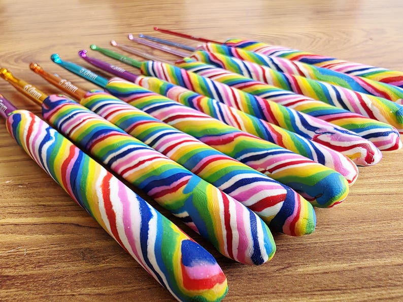 May include: A set of crochet hooks featuring metallic tips in various colours and thick, ergonomic handles with a swirling, multi-coloured rainbow pattern. Each hook displays its size clearly on the metal shaft, arranged in a row on a surface.