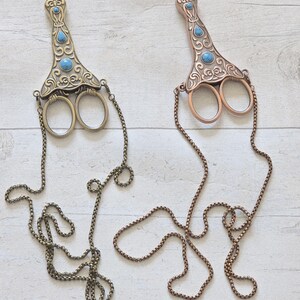 May include: Two decorative scissors with ornate handles and turquoise-coloured accents. One is bronze-toned, the other is copper-toned. Each has a chain attached. The scissors are likely for crafting or decorative purposes.