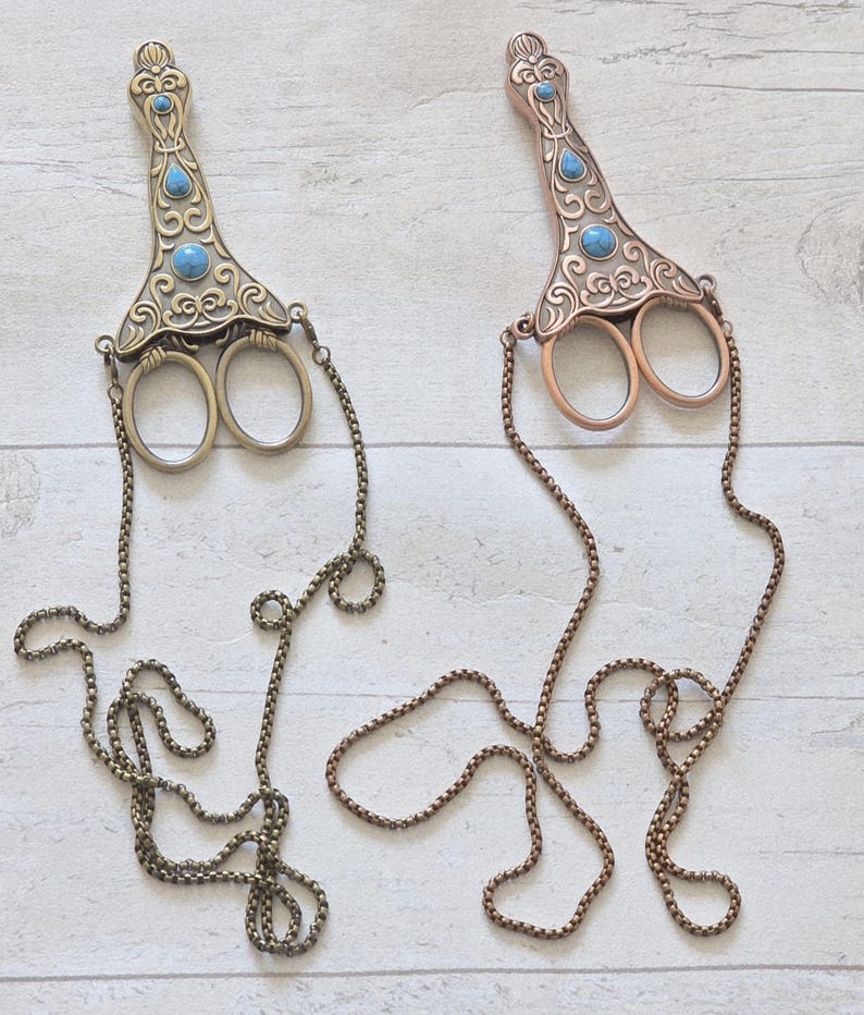 May include: Two decorative scissors with ornate handles and turquoise-coloured accents. One is bronze-toned, the other is copper-toned. Each has a chain attached. The scissors are likely for crafting or decorative purposes.