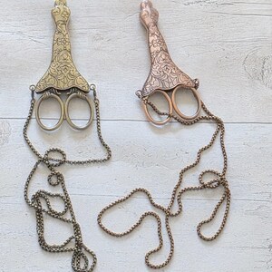 May include: Two decorative scissors with ornate handles and attached chains. One is gold-toned, the other copper-toned. The scissors have a vintage aesthetic and are displayed on a light wood-grain surface.