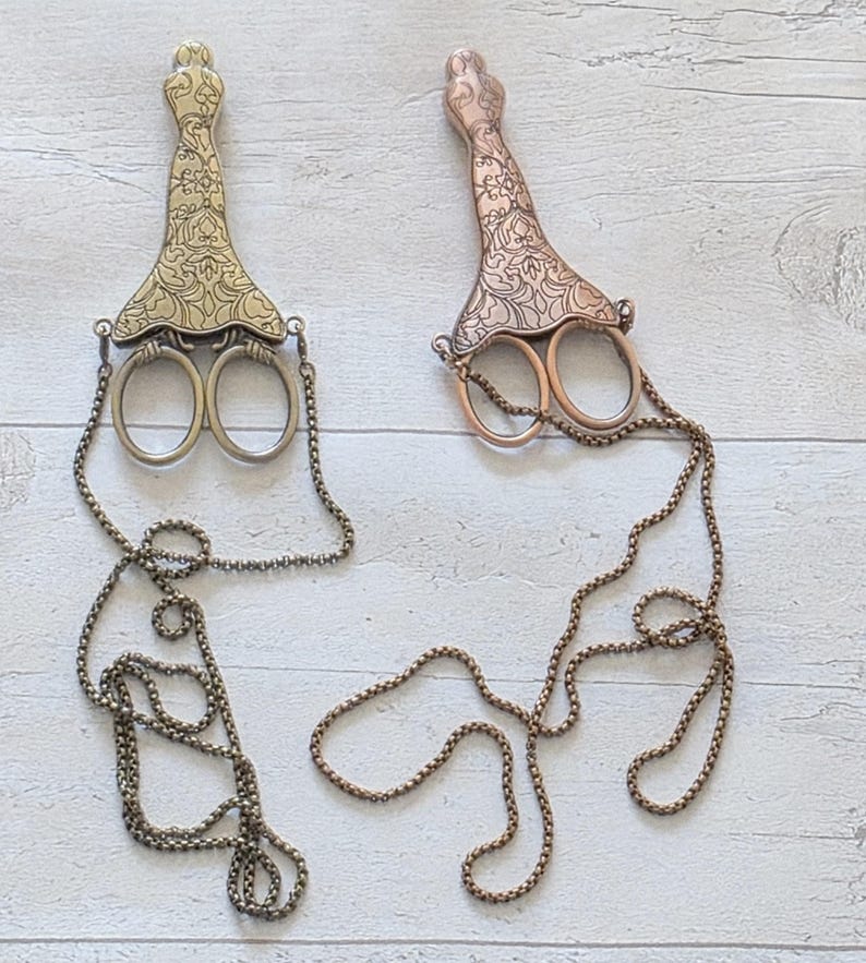 May include: Two decorative scissors with ornate handles and attached chains. One is gold-toned, the other copper-toned. The scissors have a vintage aesthetic and are displayed on a light wood-grain surface.