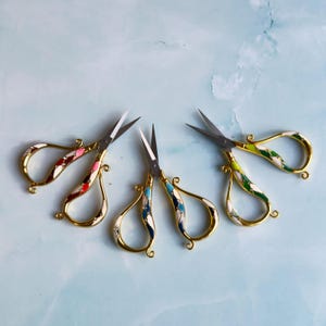 May include: Three pairs of small metal scissors with gold-toned handles featuring inlaid enamel patterns in pink, red, blue, green, and white. Each pair has sharp steel blades and ornate, curved handle designs with small decorative accents.