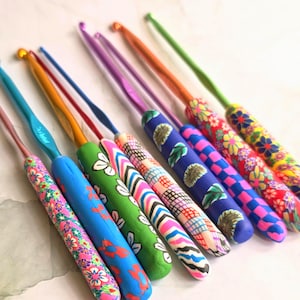 May include: A collection of nine crochet hooks with metallic shafts and handles decorated in various patterns including floral, checkered, striped, and leaf designs. The hooks feature vibrant colours such as pink, blue, green, orange, and purple.