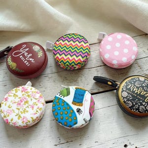 May include: Six round retractable measuring tapes with diverse patterns. One is burgundy with "Yarn Rules!" text, another has a zig-zag design, and a third is pink with white polka dots. The others feature floral, sewing-themed, and floral designs. Each tape is for measuring.