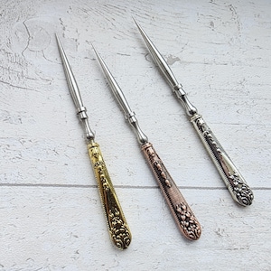 May include: Three metal awls with ornate handles. One awl has a gold handle with a floral design, another has a copper-colored handle, and the third has a silver handle. The awls have long, pointed tips.