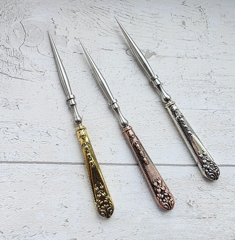 May include: Three metal awls with ornate handles. One awl has a gold handle with a floral design, another has a copper-colored handle, and the third has a silver handle. The awls have long, pointed tips.