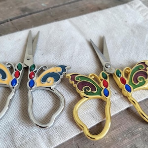 May include: Two pairs of decorative butterfly scissors. One pair has silver handles with yellow and blue wings, the other has gold handles with green and burgundy wings. Both pairs feature colourful jewel accents.