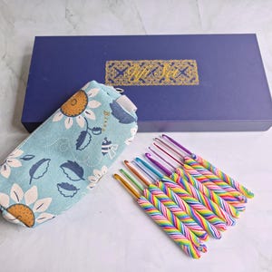 May include: A set of crochet hooks with multi-coloured striped handles arranged in a row next to a light blue fabric pouch featuring a sunflower and bee pattern, all positioned in front of a rectangular dark blue box labelled Gift Set.