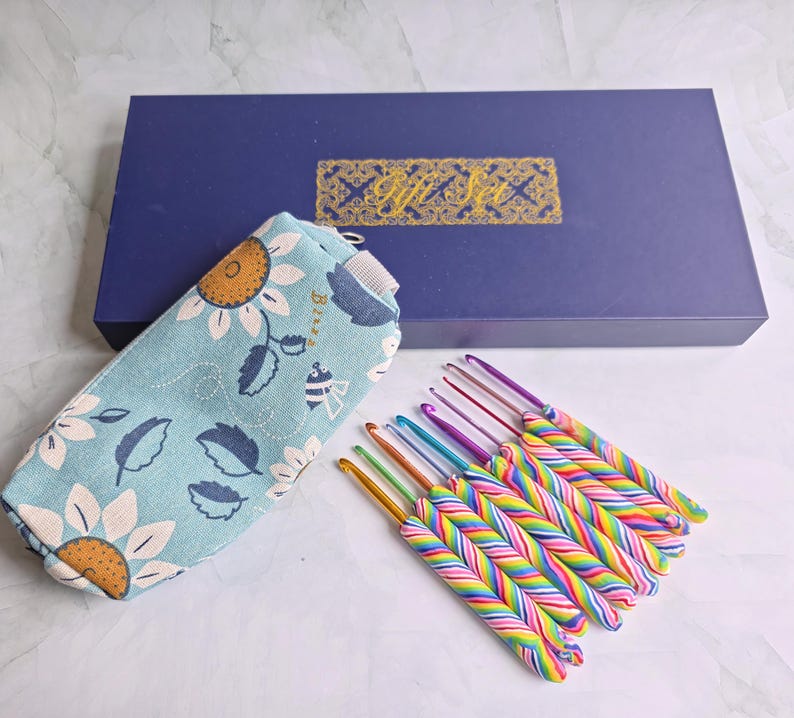 May include: A set of crochet hooks with multi-coloured striped handles arranged in a row next to a light blue fabric pouch featuring a sunflower and bee pattern, all positioned in front of a rectangular dark blue box labelled Gift Set.