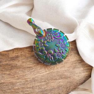 May include: Iridescent metal bottle stopper with a floral design. The stopper features a round base with embossed flowers and a handle. The colors shift from green to purple and blue. The stopper is placed on a wooden surface.