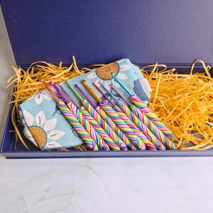 May include: A set of crochet hooks with multi-coloured striped handles arranged in a light blue fabric pouch featuring a sunflower pattern. The items are placed inside a dark blue rectangular box filled with yellow shredded paper filler.