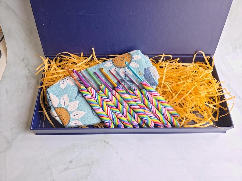 May include: A set of crochet hooks with multi-coloured striped handles arranged in a light blue fabric pouch featuring a sunflower pattern. The items are placed inside a dark blue rectangular box filled with yellow shredded paper filler.