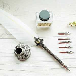 Peut inclure: Une plume d'oie blanche avec un manche en métal argenté orné, un porte-plume circulaire en métal assorti aux motifs gaufrés complexes, un encrier en verre transparent avec un bouchon octogonal noir et cinq plumes métalliques interchangeables.