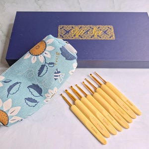 May include: A set of eight crochet hooks with light-coloured wooden handles and gold-toned metal tips, each marked with sizes ranging from 2.5mm to 4.0mm. The hooks are accompanied by a light blue fabric pouch with a sunflower print and a blue gift box.