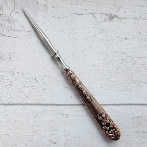 May include: A letter opener with a silver blade and a decorative handle. The handle is a copper color with floral and beaded designs. The letter opener is approximately 8 inches long.
