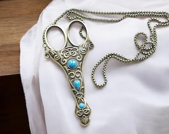 Puede incluir: Un collar de estilo antiguo con un colgante en forma de tijeras. El colgante es de color bronce con intrincados adornos y tres piedras de color turquesa. La cadena es de color bronce a juego y está sobre una tela blanca.