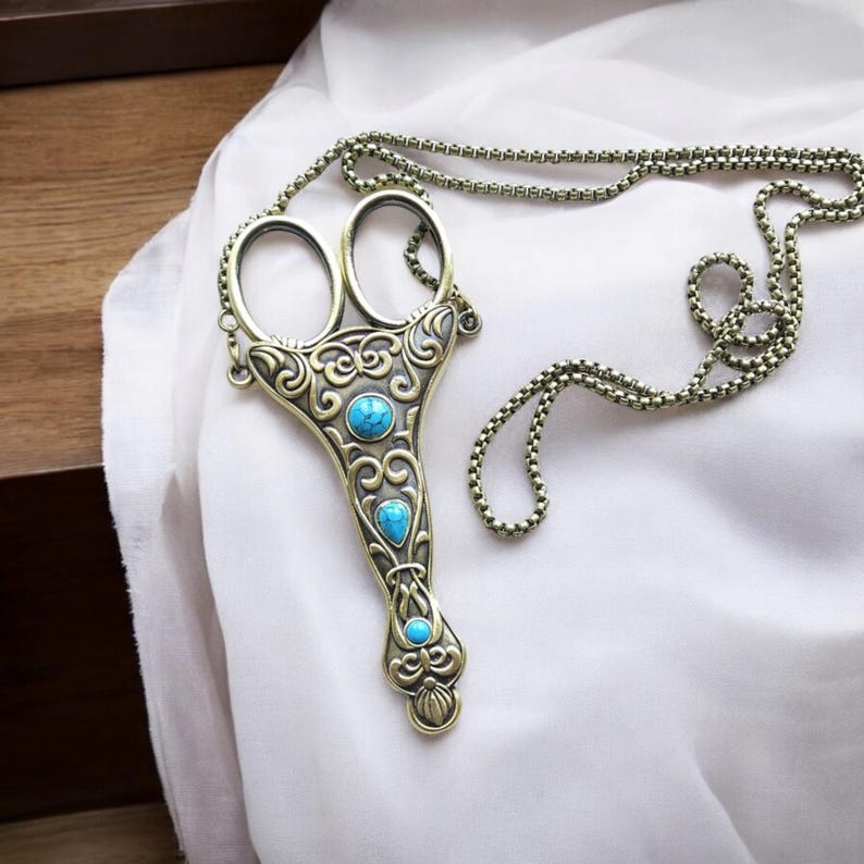 May include: A bronze-coloured necklace with a pendant in the shape of scissors. The pendant has ornate scrollwork and three turquoise-coloured stones. The chain is also bronze and rests on white fabric. The necklace is a decorative item.