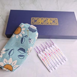 May include: A light blue fabric pouch with a sunflower and bee pattern, a dark blue rectangular gift box with the gold text Gift Set, and seven white crochet hooks with floral patterns and purple metal tips in various sizes.