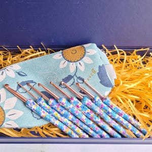 May include: A set of eight crochet hooks with rose gold metal tips and floral patterned handles, arranged in a light blue fabric pouch featuring a sunflower print, all presented inside a dark blue rectangular gift box with yellow paper filler.