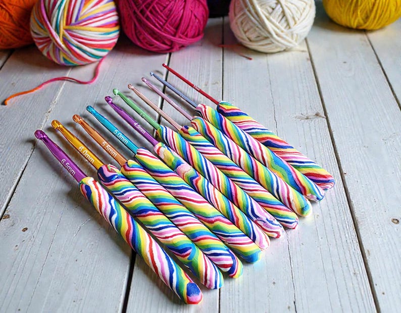 May include: A set of nine crochet hooks with colourful, swirled, rainbow-patterned handles. Each metal hook features a specific size printed on the shaft, ranging from 6.5mm down to smaller sizes, arranged in a diagonal row on a white wooden surface.