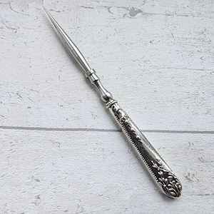 May include: A silver letter opener with a long, pointed blade and a decorative handle. The handle features floral and scrollwork designs. The opener is made of a shiny, metallic material and is set against a light-colored background.