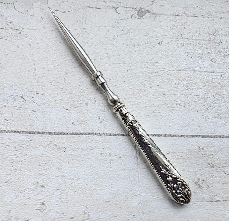 May include: A silver letter opener with a long, pointed blade and a decorative handle. The handle features floral and scrollwork designs. The opener is made of a shiny, metallic material and is set against a light-colored background.