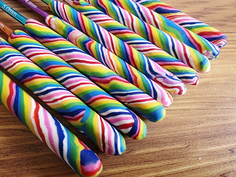 May include: A collection of crochet hooks featuring thick, custom-moulded handles with a vibrant, multi-coloured swirl pattern of red, yellow, green, blue, and pink. The metal hook tips are visible, with some showing size markings like 5.0mm.