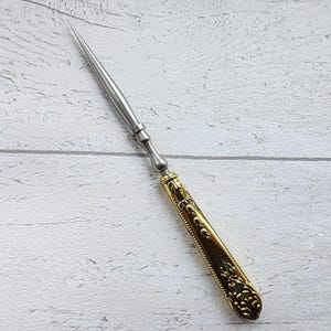 May include: A silver letter opener with a gold-colored handle. The handle features an ornate, embossed design. The blade is long and pointed, and the opener is set against a white wooden background.