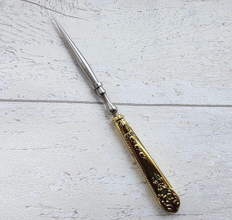 May include: A silver letter opener with a gold-colored handle. The handle features an ornate, embossed design. The blade is long and pointed, and the opener is set against a white wooden background.