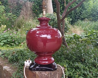 May include: A large, red ceramic table lamp with a round base and a black plug. The lamp is sitting on a stone surface in a garden setting.