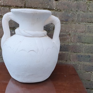 May include: A white vase with two handles and a rope-like detail around the neck. The vase has a textured surface and is set against a brick wall background. The vase is likely made of ceramic.