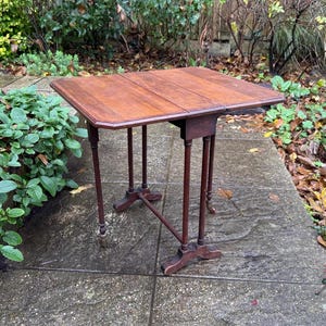 May include: A vintage wooden drop-leaf table with a rectangular top and slender legs. The table is made of dark brown wood and has a small drawer. The table's dimensions are approximately 76 cm long, 51 cm wide, and 71 cm high.