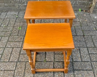 May include: Two wooden nesting tables with a warm, honey-toned finish. The tables have square tops and are supported by four legs. The smaller table fits neatly under the larger one, offering a space-saving design.
