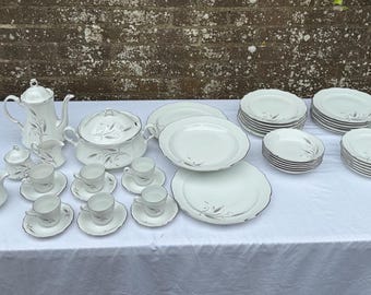 Op de afbeelding: Een compleet wit porseleinen servies met een zilveren bladeren design. De set bevat een koffiepot, serveerschalen, kopjes, schotels, kommen en borden in verschillende maten. Het servies staat op een wit tafelkleed.