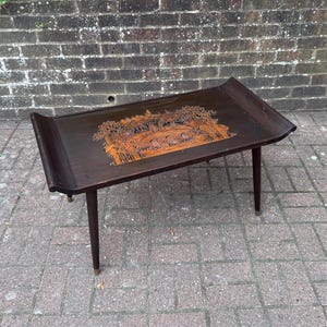 May include: A dark brown wooden coffee table with four tapered legs and curved, raised ends. The rectangular tabletop features a central inlaid design in a lighter wood tone, depicting a detailed landscape scene with trees and architectural structures.