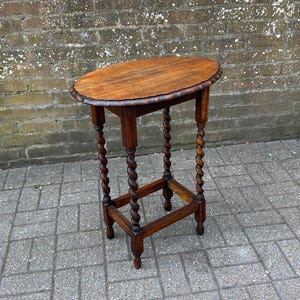 May include: A small, antique wooden side table with a dark brown finish. The oval tabletop has a decorative edge. The legs are intricately carved in a spiral design. The table is approximately 71 cm tall.